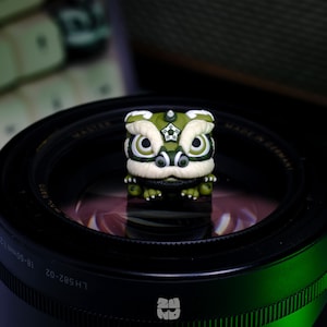 May include: A green and white lion dance head figurine with a star on its forehead. The figurine is sitting on a black camera lens.