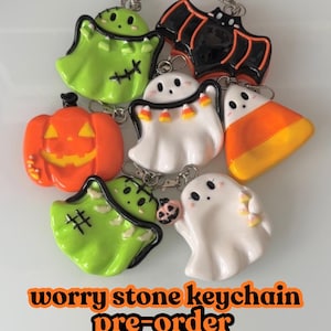 Puede incluir: Una colección de llaveros de piedra de la preocupación con temática de Halloween. Los llaveros presentan diseños como fantasmas, una calabaza, un murciélago y caramelos de maíz. Los llaveros son de varios colores, incluyendo verde, naranja, blanco y negro. También se ve el texto "worry stone keychain pre-order".