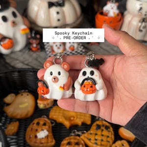 May include: Two white keychains featuring a bear and a ghost, each holding a small orange jack-o'-lantern bucket. The bear has brown ears and a candy corn, while the ghost has a black bat on top. Text on the image reads Spooky Keychain PRE-ORDER.