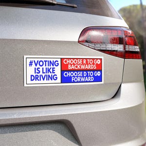 Op de afbeelding: Een rechthoekige bumpersticker met de tekst "#VOTING IS LIKE DRIVING" in blauw en wit, en "CHOOSE R TO GO BACKWARDS" in rood, en "CHOOSE D TO GO FORWARD" in blauw. De sticker is bevestigd aan de achterkant van een zilveren auto.