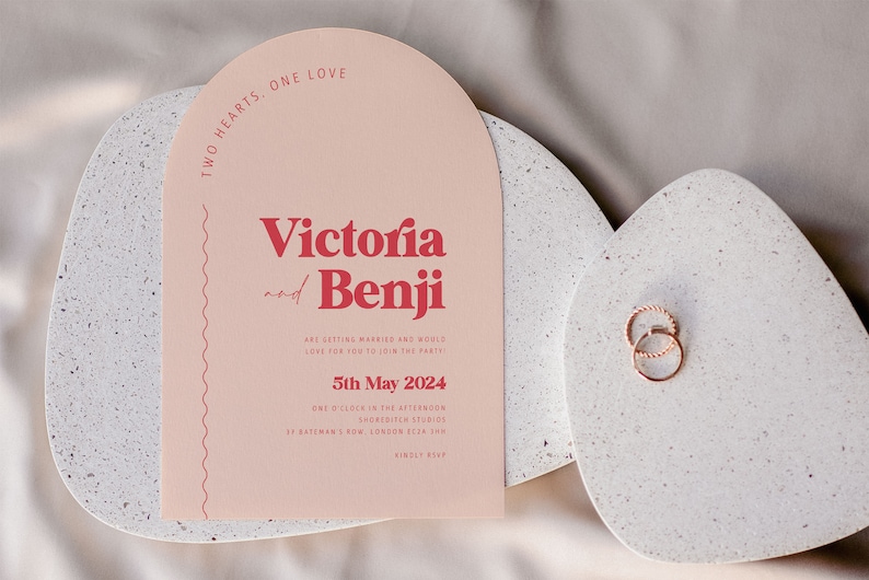 May include: A pink wedding invitation with the text "Two Hearts, One Love" at the top. The names "Victoria and Benji" are in red text. The invitation includes the date "5th May 2024" and the location "Shoreditch Studios, 37 Bateman's Row, London EC2A 3HN".