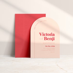 May include: A peach-colored wedding invitation with a rounded arch shape. The invitation features the text "Two Hearts, One Love" in a red font. The names "Victoria and Benji" are in a larger red font. The date "5th May 2024" is also in red font.