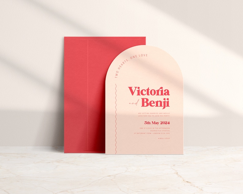 May include: A peach-colored wedding invitation with a rounded arch shape. The invitation features the text "Two Hearts, One Love" in a red font. The names "Victoria and Benji" are in a larger red font. The date "5th May 2024" is also in red font.