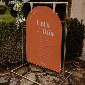 以下が含まれることがあります： 桃色のアーチ型の看板に、白い文字で「Let's do this」と書かれています。看板には、結婚式当日のスケジュールが記載されており、式典、飲み物、カナッペ、夕食の呼びかけ、スピーチ、食事、甘いもの、ケーキカット、ダンス、軽食、馬車などが含まれています。看板は金色の金属製のスタンドに掛けられています。