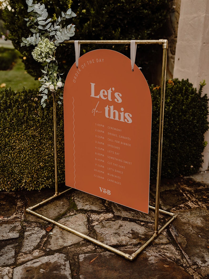 以下が含まれることがあります： 桃色のアーチ型の看板に、白い文字で「Let's do this」と書かれています。看板には、結婚式当日のスケジュールが記載されており、式典、飲み物、カナッペ、夕食の呼びかけ、スピーチ、食事、甘いもの、ケーキカット、ダンス、軽食、馬車などが含まれています。看板は金色の金属製のスタンドに掛けられています。