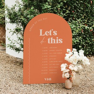 以下が含まれることがあります： ピーチ色のアーチ型の看板に、「Order of the Day」と「Let's do this」という白い文字が書かれています。看板には、結婚式のリセプションのイベントの順番が書かれており、式典、飲み物、夕食、スピーチなどが含まれています。