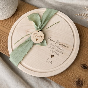 May include: A round wooden serving board with a light green ribbon and a small wooden tag that says "Danke" with a heart. The board has the text "Kleine Menschen brauchen so gro&szlig;e Herzen wie deins" and the name "Nils" engraved on it.