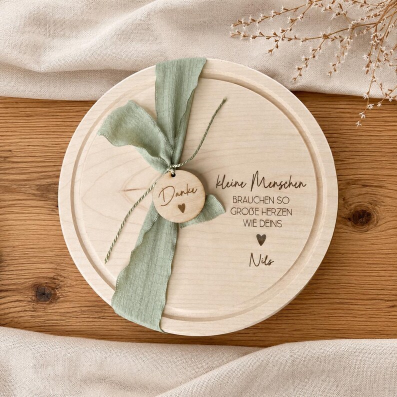May include: A round wooden serving board with a light green ribbon and a small wooden tag that says "Danke". The board has the text "kleine Menschen brauchen so gro&szlig;e Herzen wie deins Nils".