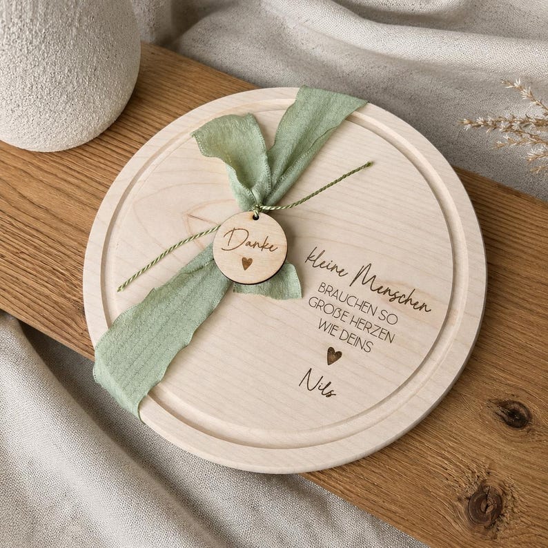 May include: A round wooden serving board with a light green ribbon and a small wooden tag that says "Danke" with a heart. The board has the text "Kleine Menschen brauchen so gro&szlig;e Herzen wie deins" and the name "Nils" engraved on it.