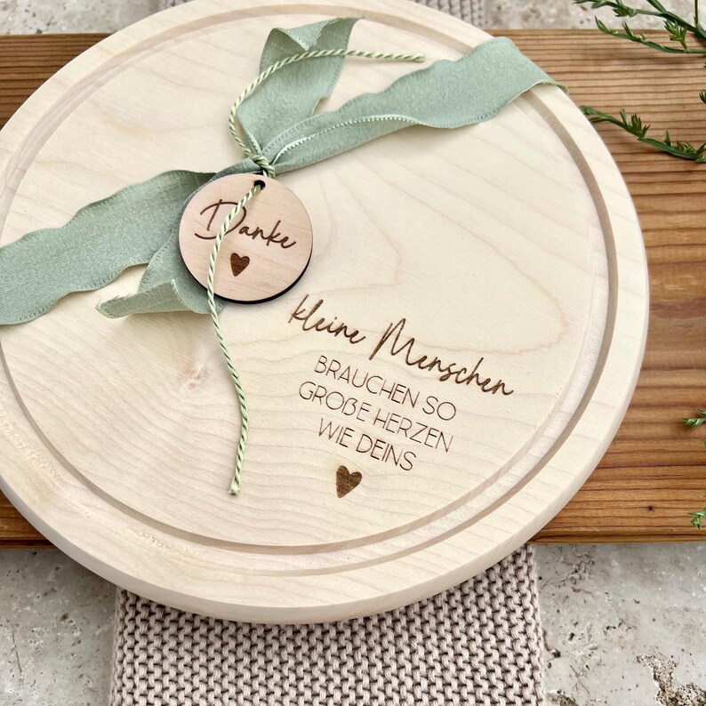May include: A round wooden serving board with a decorative edge. The board has engraved text and a small wooden tag with the word "Danke" and a heart. A green ribbon and string are attached.
