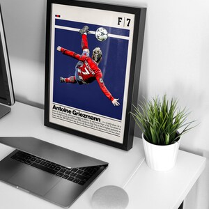 May include: A framed poster featuring a soccer player in mid-air, kicking a soccer ball. The poster is blue and white with the text "F/7 Antoine Griezmann" at the top. The player is wearing a red and white jersey with the number 7 on it.
