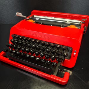 May include: A vibrant red Valentine typewriter with black keys and the brand name visible. The keys are arranged in a QWERTY layout. The typewriter is on a black surface.