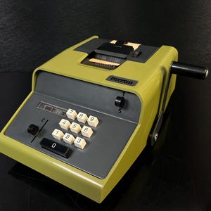 Op de afbeelding: Vintage olijfgroene rekenmachine met een grijs toetsenbord en witte cijfertoetsen. De rekenmachine heeft een zwarte hendel en een papieruitvoersleuf. De merknaam is zichtbaar aan de bovenkant. Een klassiek kantoorbenodigdheid.