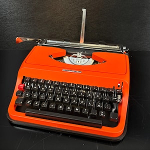 May include: A bright orange Underwood 30 typewriter featuring a QWERTY keyboard with black keys, a single red key on the left side, a metal carriage, and a paper support arm extending upward from the back of the machine's main body.