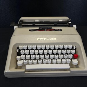 May include: A vintage Olivetti Lettera 35 typewriter in a light beige color. The keyboard features white keys with black lettering, a red key on the right, and a white space bar. The model name is visible on the front.
