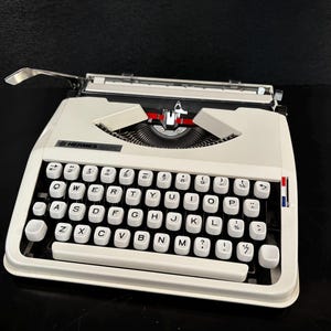 Può includere: Una macchina da scrivere Hermes 3000 bianca vintage con tasti neri e una leva di ritorno del carrello cromata. La tastiera presenta un layout QWERTY. La macchina da scrivere ha un accento rosso, bianco e blu sul lato destro.