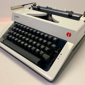 Könnte beinhalten: Eine weiße Olympia Monica Schreibmaschine mit schwarzer Tastatur und rotem Logo. Die Schreibmaschine hat einen Wagenrücklaufhebel und einen Papierfreigabehebel.