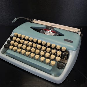 Puede incluir: Una máquina de escribir Triumph vintage en una combinación de colores azul claro y gris. La máquina de escribir presenta teclas redondas de color blanquecino con letras negras, un soporte de papel cromado y la marca "Triumph" en la parte superior. Las teclas están dispuestas en un diseño QWERTY estándar.