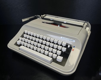 May include: A vintage gray typewriter with white keys and a silver carriage return lever. The typewriter has a classic design with a full keyboard and a paper feed mechanism. The brand name is visible on the front of the typewriter.