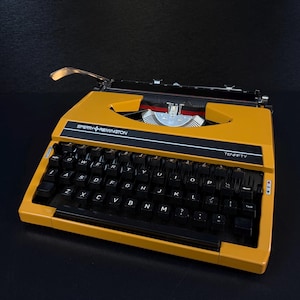 Könnte beinhalten: Eine senfgelbe Sperry Remington Ten-Fifty tragbare Schreibmaschine mit schwarzer Tastatur und weißer Beschriftung, einem Wagenrücklaufhebel aus Metall und einer schwarzen Farbbandabdeckung. Markenname und Modell sind weiß auf der Vorderseite.
