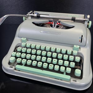 Può includere: Una macchina da scrivere Hermes vintage in grigio chiaro con tasti e dettagli verde menta. La tastiera presenta un layout QWERTY standard. La parola "HERMES" è visibile sulla parte superiore della macchina da scrivere.