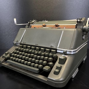 Op de afbeelding: Een olijfgroene vintage schrijfmachine met een volledig toetsenbord en een papierrol. De schrijfmachine heeft een metalen frame met zilveren afwerking en donkergroene toetsen. Het ontwerp is klassiek en functioneel.