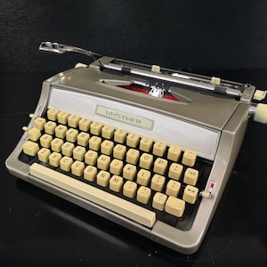 May include: A vintage Brother typewriter in silver and cream. The typewriter features a full keyboard with cream-colored keys, a silver carriage, and the brand name "BROTHER" displayed prominently. The design is classic and retro.