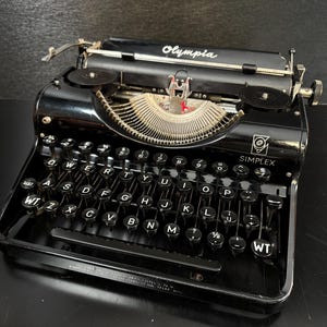 May include: A black Olympia Simplex typewriter featuring a QWERTY keyboard layout with circular black keys, white lettering, two ribbon spools, and a metal typebar segment. The brand name Olympia is printed in white on the top casing.