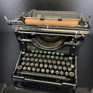 Puede incluir: Máquina de escribir Underwood negra con teclas verdes y blancas. La máquina de escribir tiene un rodillo marrón y la palabra "Underwood" es visible en la parte superior. Las teclas están dispuestas en un diseño QWERTY.