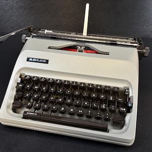 May include: A light gray Adler typewriter featuring a QWERTY keyboard with black keys, a metal carriage, and a paper support arm. The brand name Adler is printed in black on a white rectangular label on the front left side of the machine casing.
