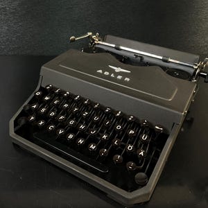 Könnte beinhalten: Eine schwarze Adler-Schreibmaschine mit weißen Buchstaben. Das Wort "ADLER" ist auf der Vorderseite aufgedruckt. Die Schreibmaschine hat einen silbernen Wagenrücklaufhebel und ein schwarzes Gehäuse. Die Tasten sind in QWERTZ angeordnet.