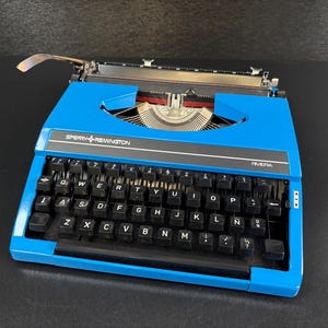 Può includere: Una macchina da scrivere Sperry Remington Riviera blu brillante dotata di una tastiera QWERTY nera con lettere bianche. Il dispositivo include una leva di ritorno del carrello in metallo sul lato sinistro e un pannello nero con il marchio.