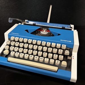 May include: A vintage Olympia Traveller typewriter in blue and white. The typewriter has a full keyboard with white keys and black lettering. The carriage return lever is silver. The word "Traveller" is printed on the front.