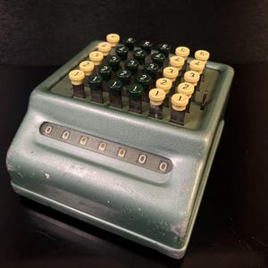 Op de afbeelding: Vintage groene mechanische rekenmachine met gele en groene cijfertoetsen. De rekenmachine heeft een reeks cijferwielen met nullen. De toetsen zijn gerangschikt in rijen en kolommen, met cijfers op elke toets.