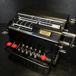 May include: A vintage black Facit mechanical calculator with red buttons and a crank handle. The calculator has a series of number keys and display windows. The Facit logo is visible on the front.
