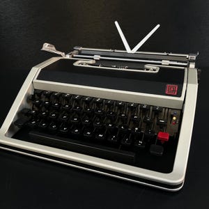 May include: A portable typewriter with a silver-toned frame and a black keyboard. The device features a red key on the right side, a black top panel with a red logo, and two white paper guides extending upwards from the rear carriage assembly.
