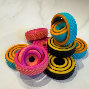 May include: A selection of brightly coloured 3D-printed fidget toys. The toys feature various designs, including spheres, rings, and interlocking shapes. Colours include blue, orange, pink, yellow, and black. The toys have textured surfaces.