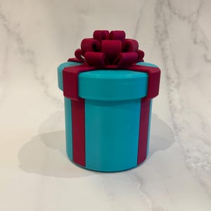 May include: A cylindrical gift box in a vibrant turquoise color, adorned with a deep magenta ribbon and bow. The box has a round lid and vertical magenta stripes. The bow is intricately designed, adding a decorative touch to the present.