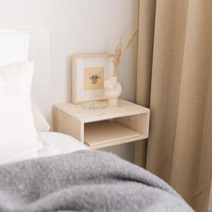 Op de afbeelding: Een lichtgekleurd zwevend nachtkastje van hout met een ingelijste bijenillustratie, een witte vaas met gedroogde bloemen en een klein glazen schaaltje. Het nachtkastje staat naast een bed met een grijze deken en witte kussens.