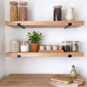May include: Wooden shelves with various kitchen items. Glass jars filled with pasta, grains, and coffee beans. A small potted plant, spice jars, and a ceramic pitcher are also visible. The shelves are supported by black metal brackets.
