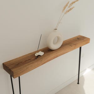 Könnte beinhalten: Ein Holztisch mit natürlicher Holzoberfläche, getragen von schwarzen Metallbeinen. Auf dem Tisch steht eine weiße Keramikvase mit kreisförmigem Design und getrockneten Pflanzen. Ein Räucherstäbchen und ein wolkenförmiger Halter sind ebenfalls vorhanden.