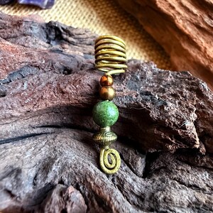 Könnte beinhalten: Ein goldfarbenes Haaraccessoire mit Spiraldesign und grünen und braunen Perlen. Der Haarschmuck ist aus Metall gefertigt und hat einen gewickelten Aufsatz. Die Perlen sind rund und haben eine strukturierte Optik. Das Accessoire wird auf einem Holzstück präsentiert.