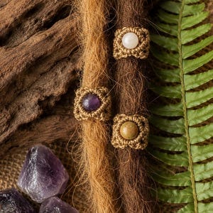 Könnte beinhalten: Drei Haarperlen mit quadratischem, beigefarbenem Makramee-Garn, das um zentrale Cabochon-Steine gewebt ist. Eine Perle enthält einen weißen Stein, eine einen violetten Amethyst und eine einen gesprenkelten grünen Stein.