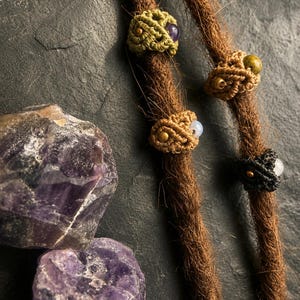 Può includere: Quattro perline per capelli in macramè color verde oliva, beige e nero, ognuna con una piccola pietra preziosa e una minuscola perlina metallica. Le perline sono infilate su due dreadlock marroni, accanto a due grandi ametiste grezze.