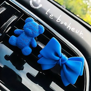 May include: Two blue car air fresheners, one shaped like a teddy bear with the word "Smile" repeated on it, and the other shaped like a bow. The text "Le bonheur" is written on the black surface behind the air fresheners.