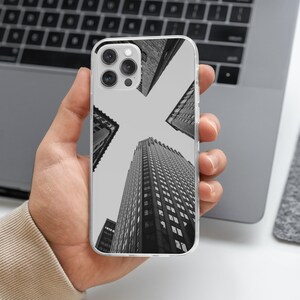 Peut inclure: Coque de téléphone transparente avec une image en noir et blanc de gratte-ciels vus d'en bas. Les bâtiments convergent vers le centre, créant une perspective unique. Le téléphone est tenu dans une main.