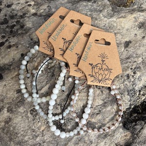May include: Four white moonstone beaded bracelets with brown cord and a brown paper tag that says "Handskien" and "Made by Athumi".