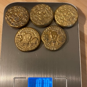 Puede incluir: Una colección de seis monedas doradas con diseños de calaveras y huesos cruzados, dispuestas sobre una báscula de cocina digital. La báscula muestra un peso de 0,485 kg. Las monedas tienen detalles intrincados y un aspecto envejecido.