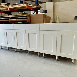 May include: A set of six white kitchen cabinet doors with raised panels and a simple design. The doors are mounted on adjustable legs and are ready to be installed.