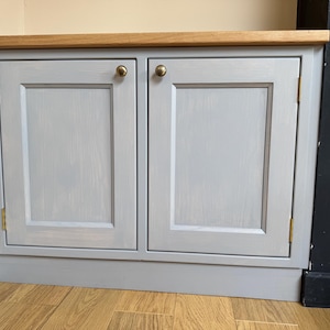 May include: A two-door cabinet painted in light grey, with a natural wood top and brass-coloured knobs and hinges. The cabinet is set against a light beige wall and a dark grey wall.